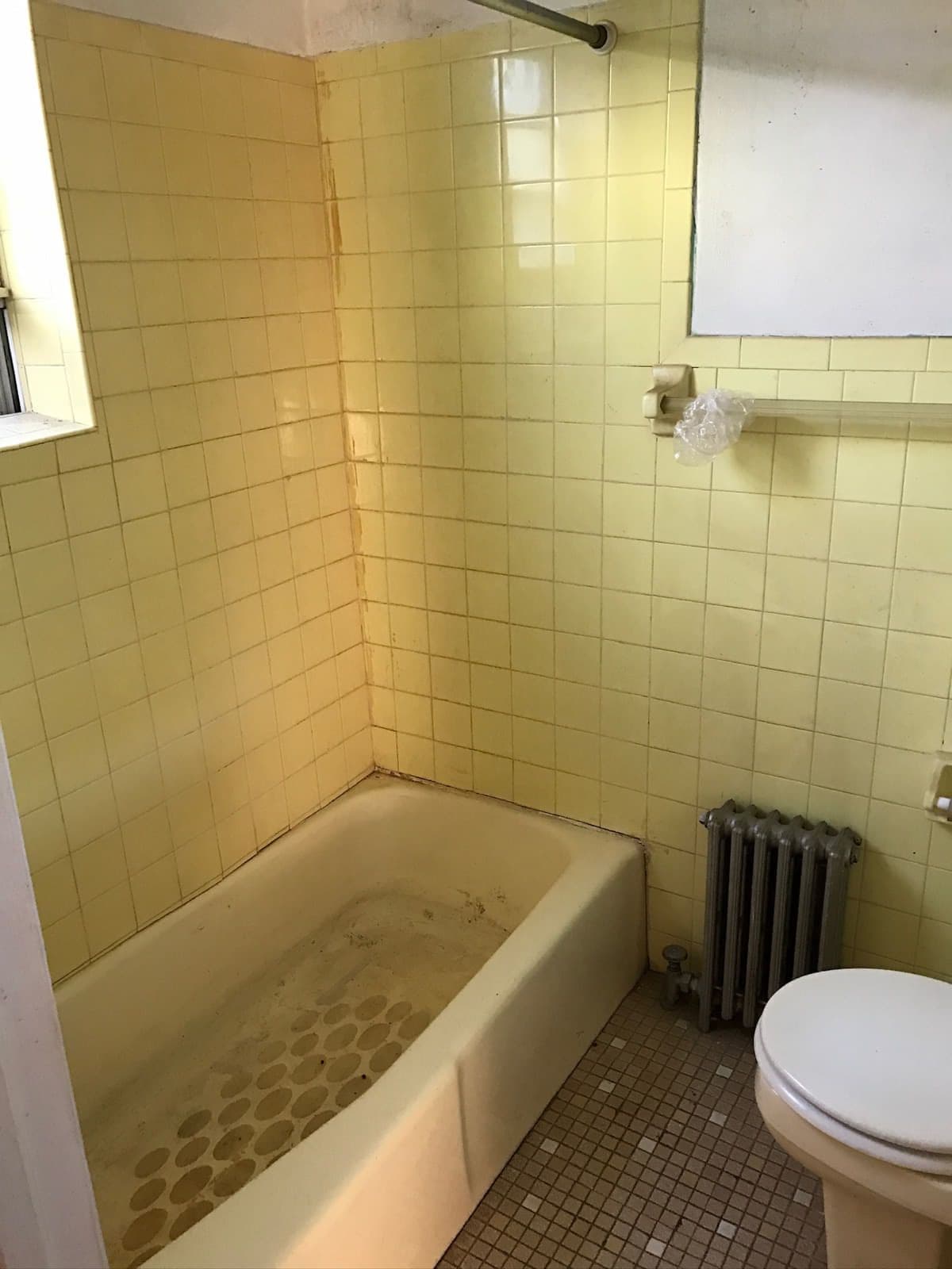 The original bathroom: yellow wall tile floor to ceiling, a stained cast-iron tub, a mosaic tile floor and a radiator beside the toilet.