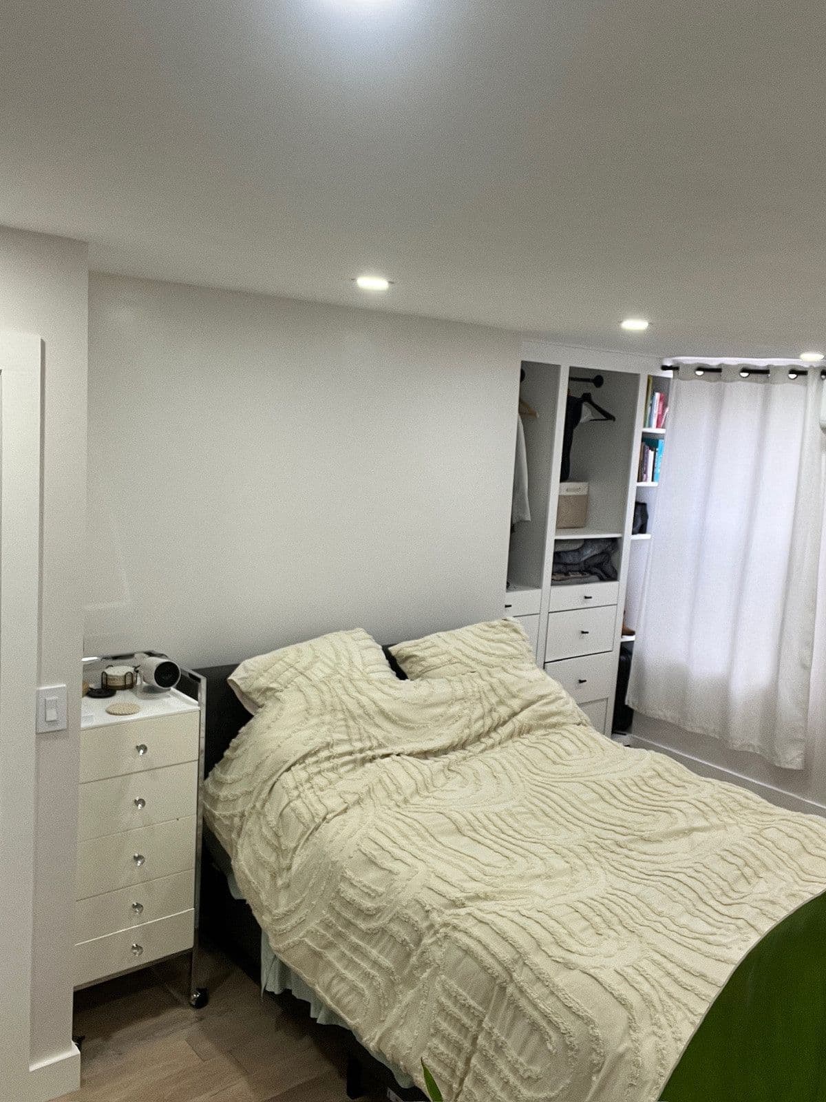The finished bedroom with recessed ceiling lights, wood-look tile floor, fitted white storage and a made bed.