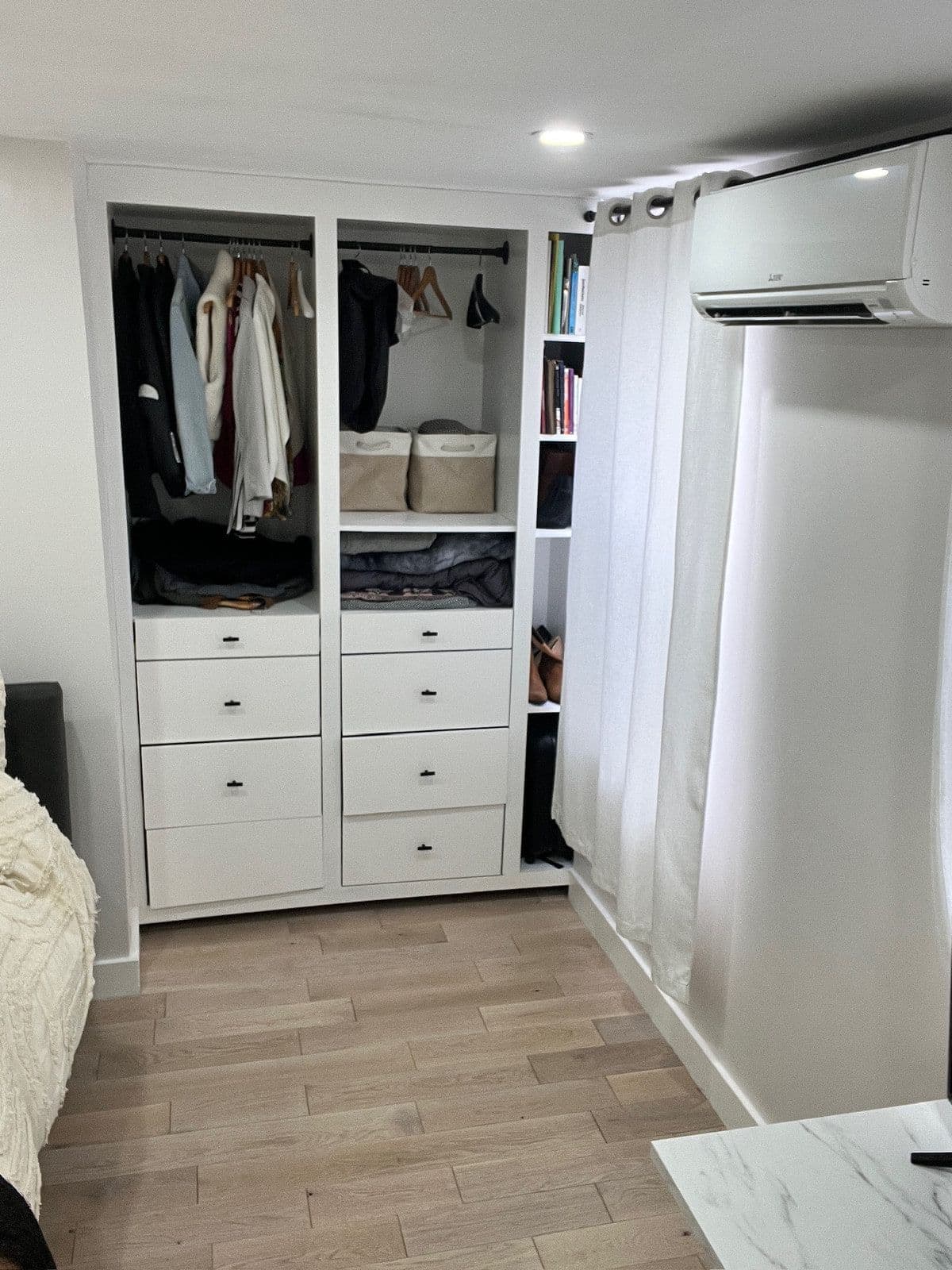 The same wall rebuilt as a floor-to-ceiling white closet system — hanging rails, banks of drawers and open shelving — beside a wall-mounted air conditioner.