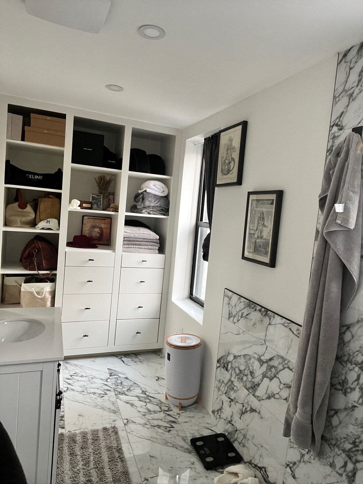 The finished bathroom from the same corner: a white vanity with a black tap, built-in shelving and cupboards, and marble tile on every wall.