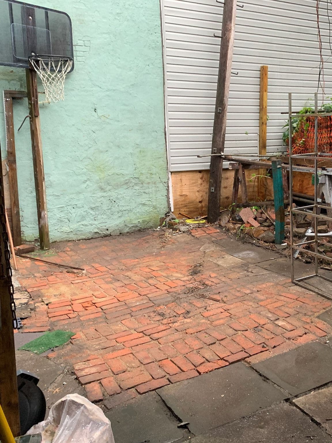 The back yard before work started: a basketball hoop on a painted wall, cracked brick and flagstone paving, a chain-link fence, and stacked scrap and shelving under a lean-to roof.