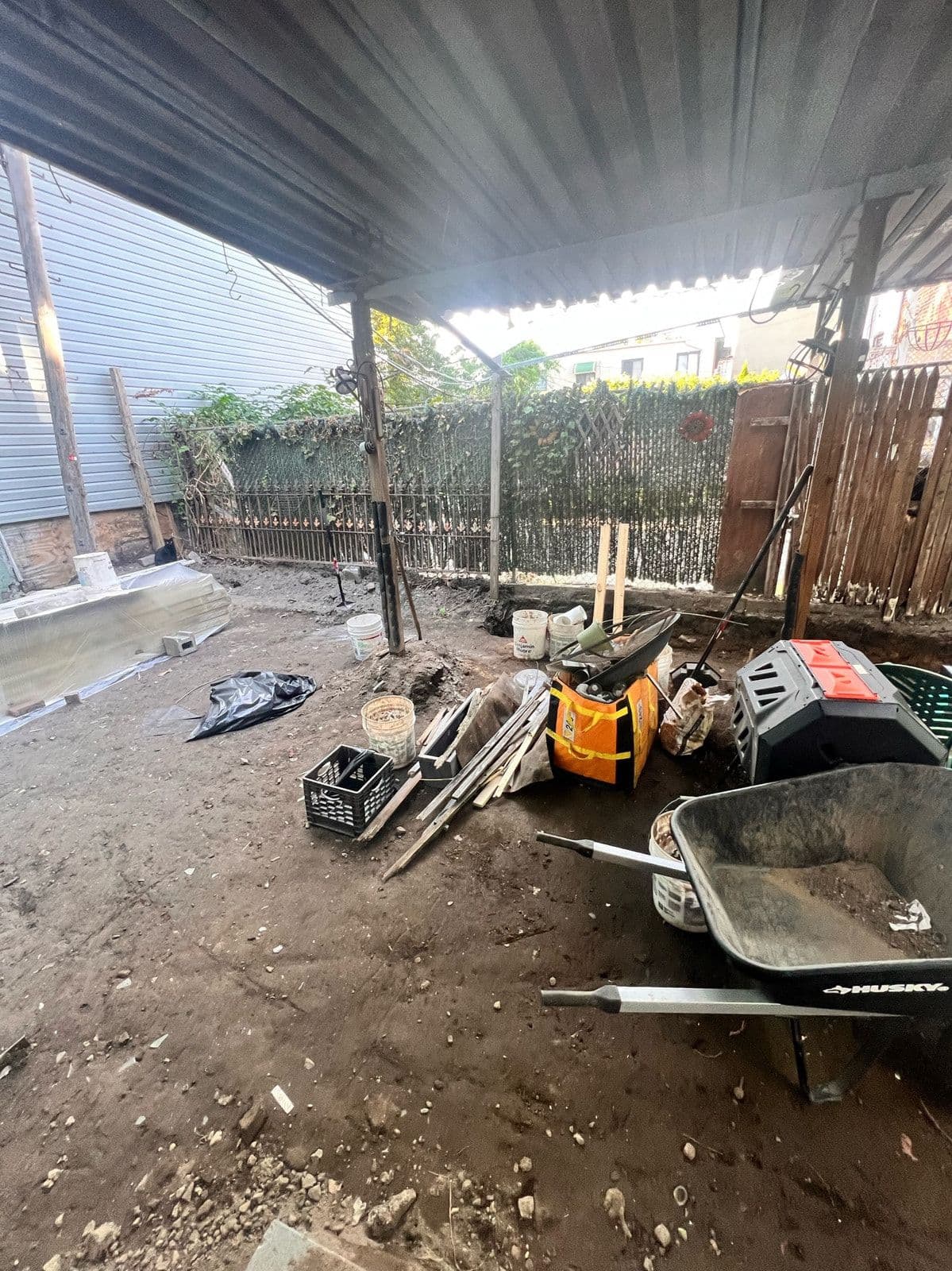 The yard dug out to bare earth with the old lean-to roof still overhead, a wheelbarrow and a plate compactor parked in the middle.